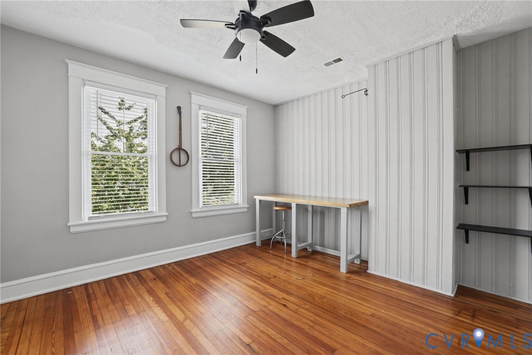 Hardwood flooring, two windows with blinds, ceiling fan, accent wall with vertical paneling, and built-in shelving