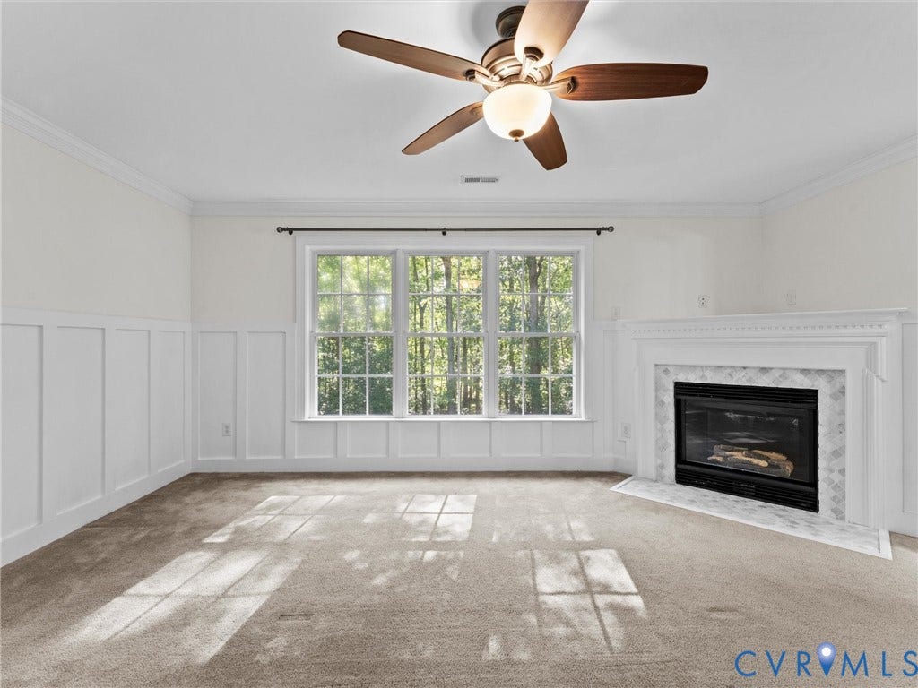Carpeted room featuring a ceiling fan with integrated lighting, crown molding, board and batten wall detailing, a three-pane window, and a corner fireplace with a tiled surround