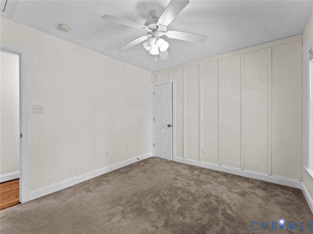 Neutral tone wall paint, textured carpeting, white baseboards, ceiling fan with light fixture, decorative wall paneling