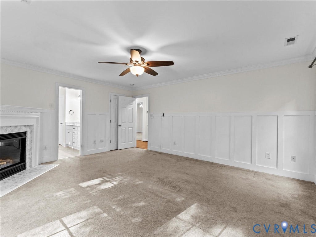 Spacious room featuring carpeting, a ceiling fan, a fireplace with a white mantel and marble-look surround, and wainscoting along the walls