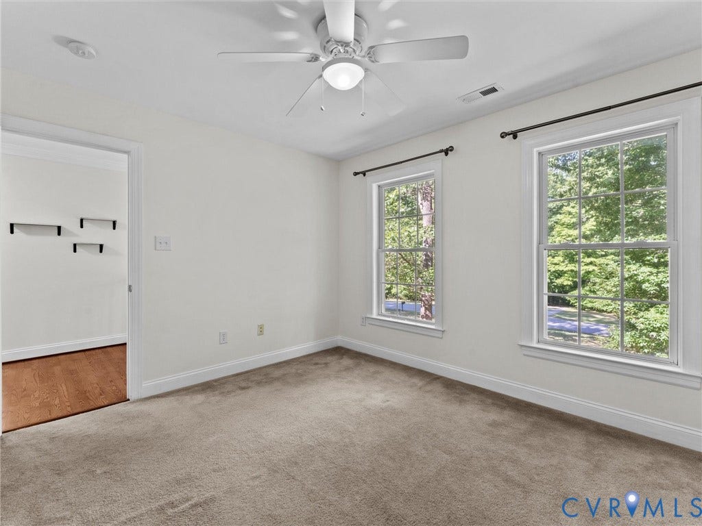 Carpeted interior space with a ceiling fan, two windows, and an adjacent room with wood-finish flooring