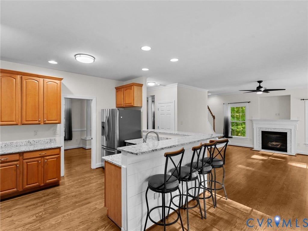Open-concept kitchen and living area featuring wood-finish flooring, wood cabinetry, stainless steel refrigerator, white granite countertops, and a fireplace with a white mantel