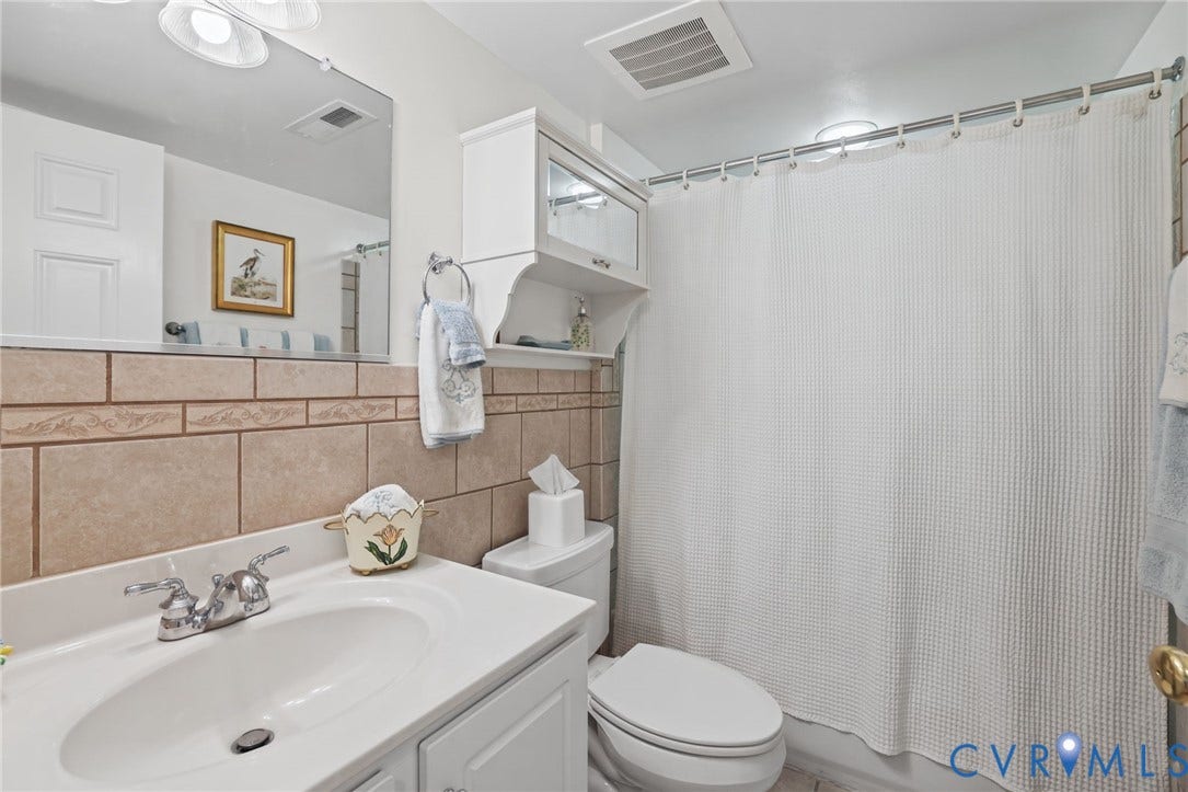 Bathroom featuring a white vanity with an integrated oval sink, chrome dual-handle faucet, large wall-mounted mirror, neutral stone-finish wall tile, and white cabinetry