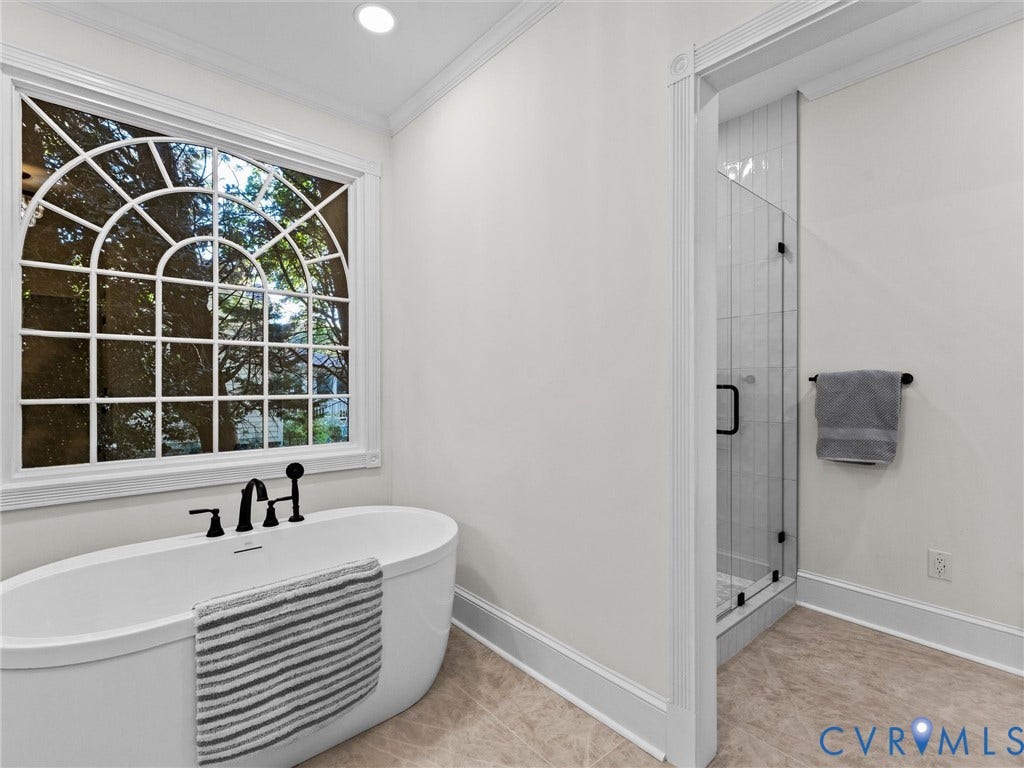 Freestanding soaking tub with matte black fixtures, arched window with intricate grid pattern, glass-enclosed shower with tile surround, and tile flooring