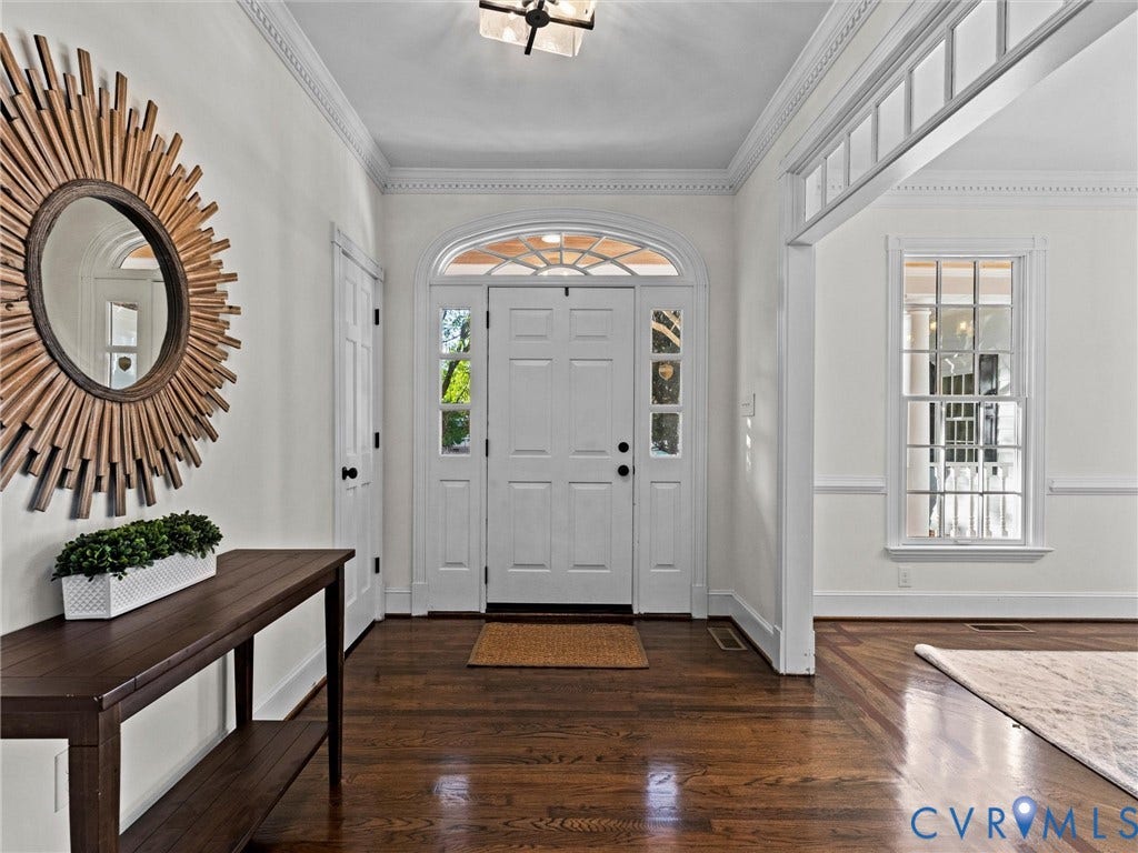 Elegant foyer featuring rich hardwood flooring, white six-panel double doors with sidelights and transom window, decorative sunburst mirror, and wainscoting details