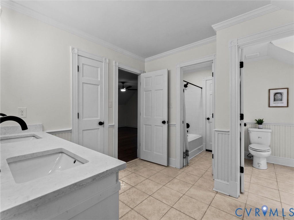 Dual-sink vanity with light-toned countertop, tiled flooring, wainscoting, and crown molding
