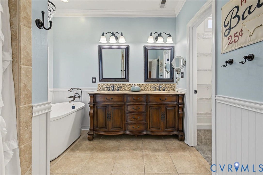 Dual-sink vanity with wood-finish cabinetry, freestanding soaking tub, ceramic tile flooring, beadboard wainscoting, and tile backsplashes