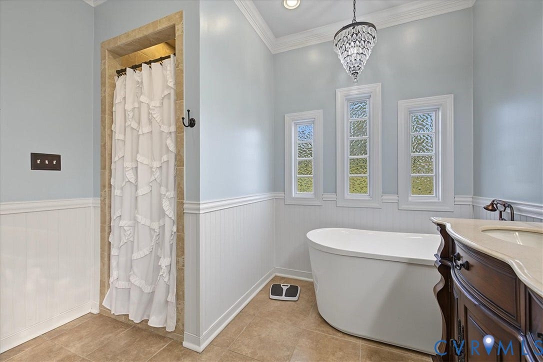 Freestanding soaking tub, stone-finish tile flooring, beadboard wainscoting, crystal-style chandelier, and tiled shower surround