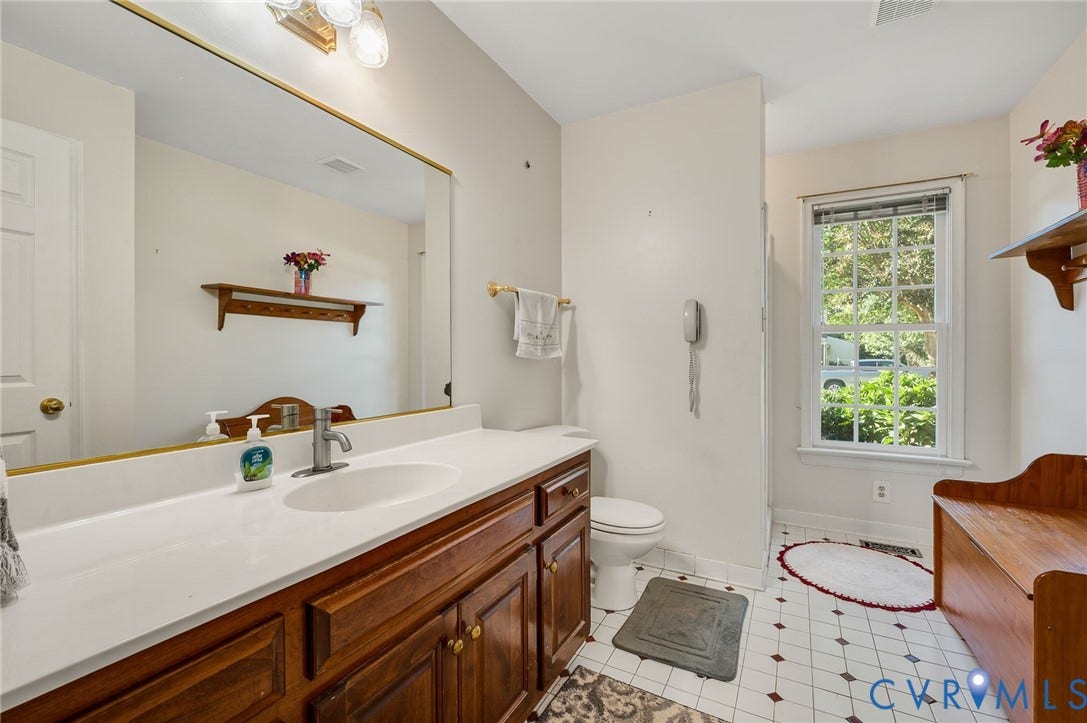 Vanity with a white countertop and wood-finish cabinetry, a large wall-mounted mirror, white toilet, and a window with blinds