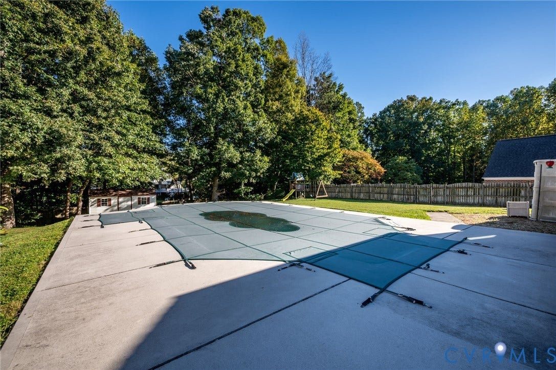 Expansive concrete patio surrounding a covered swimming pool, featuring mature trees and a wood fence