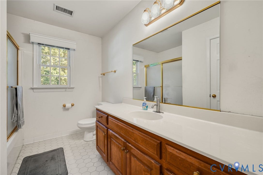 White hexagonal tile flooring, wood-finish vanity with ample storage, integrated white countertop, large wall-mounted mirror, and a window with natural light