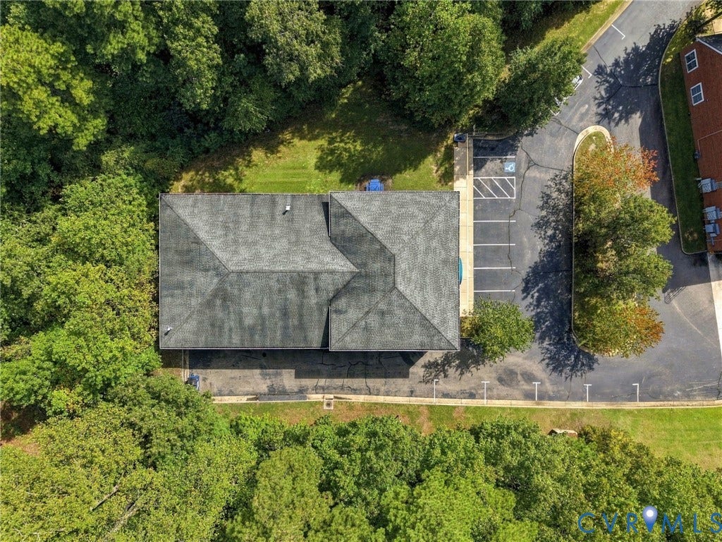 Gabled roof structure surrounded by mature trees, featuring a paved parking area with marked spaces and a building exterior