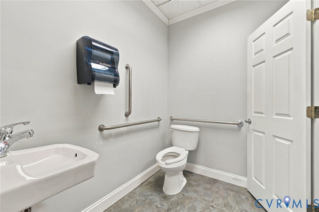 White paneled door with brass-toned hinges, wall-mounted sink with chrome faucet, toilet with white base, and durable flooring
