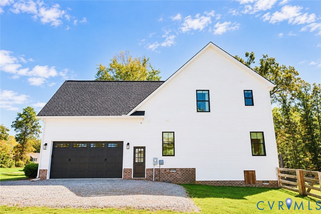 6265 Bushnell Drive, Rochambeau Estates, New Kent, VA 23124 White siding exterior with a dark shingle roof, integrated two-car garage, brick foundation detailing, and black-framed windows