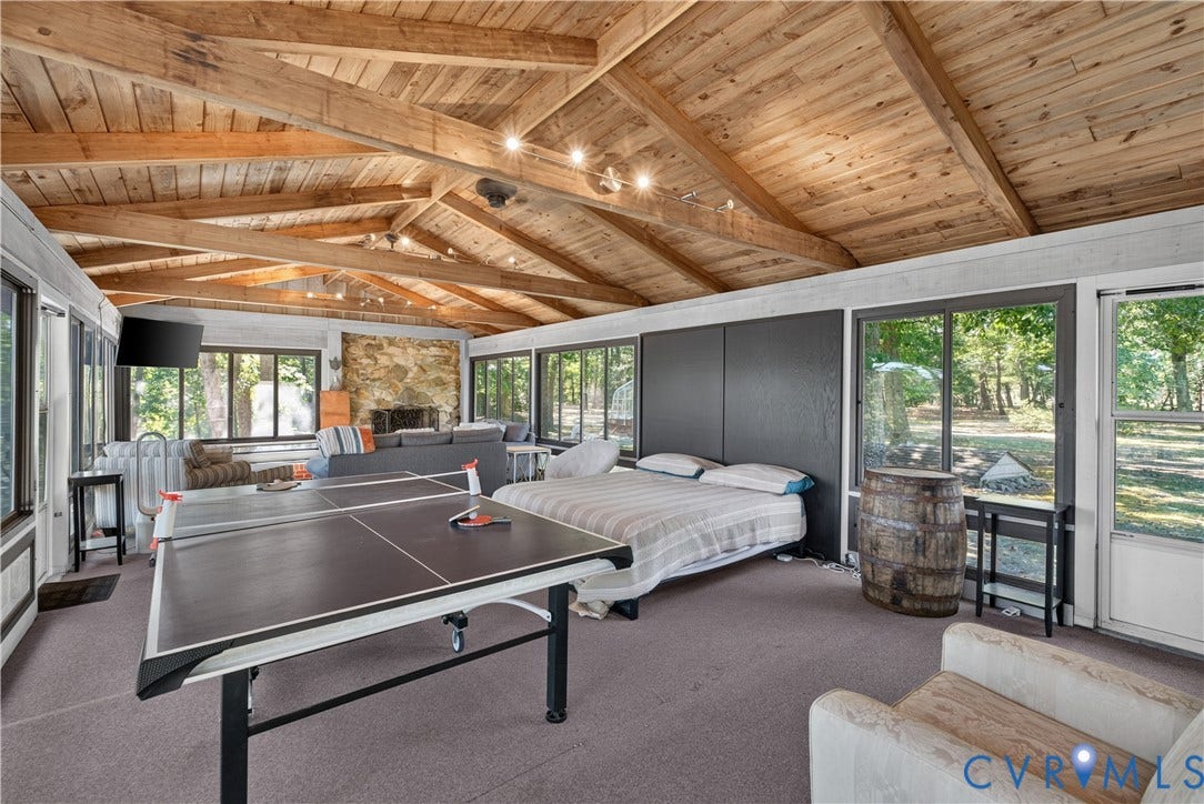 Vaulted wood-finish ceiling with exposed beams, stone-finish fireplace, multiple large windows, and wall-to-wall carpeting