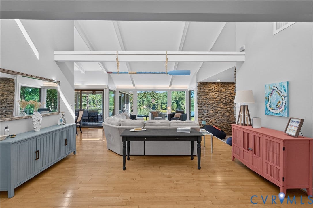 Vaulted ceilings with exposed beams, wood-finish flooring, a prominent stone accent wall, and expansive windows