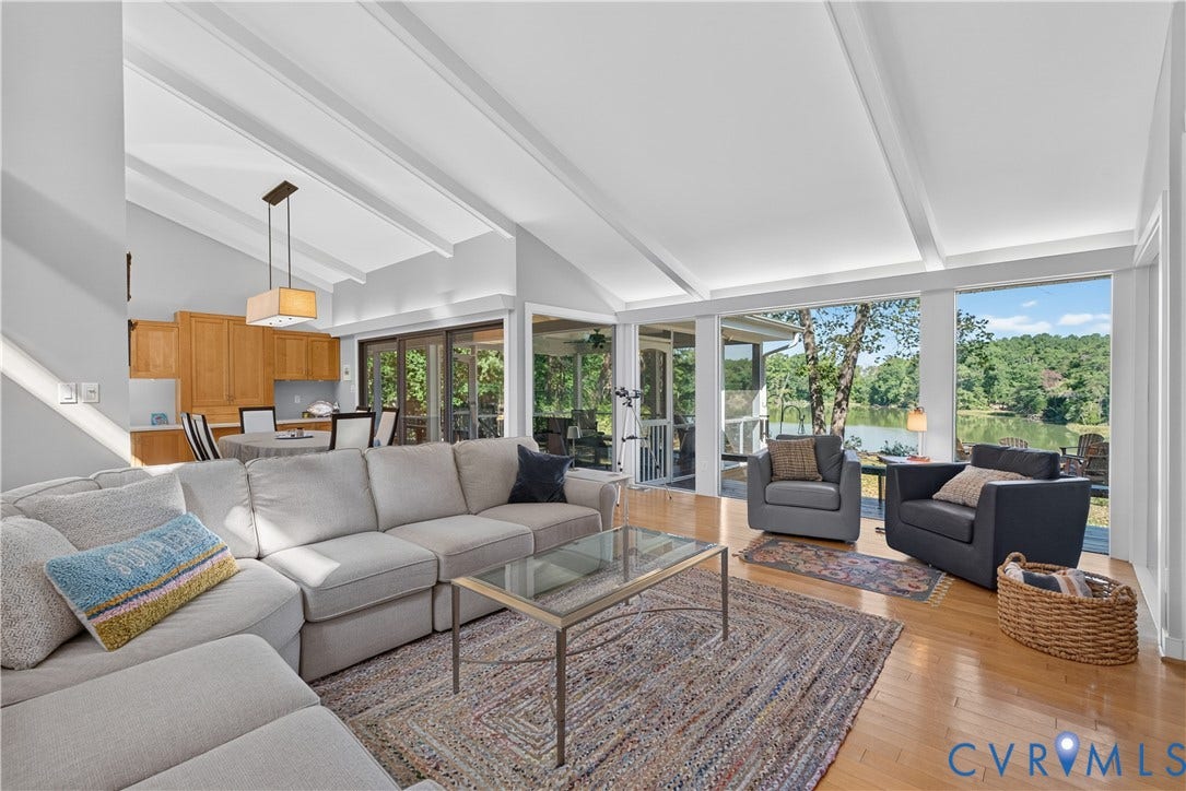 Vaulted ceilings with exposed beams, wood-finish flooring, a contemporary chandelier, and expansive windows showcasing a prominent body of water