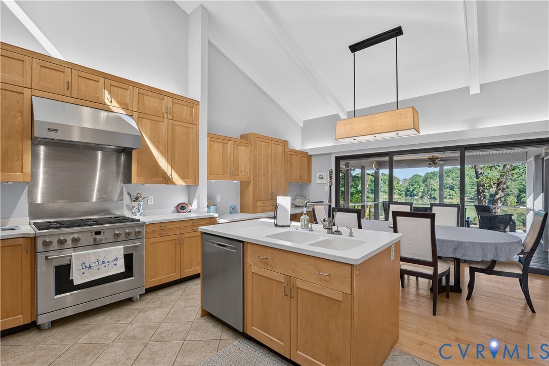 Vaulted ceiling with exposed beams, wood cabinetry, stainless steel range with hood, center island with dual sink, and sliding glass doors to outdoor area