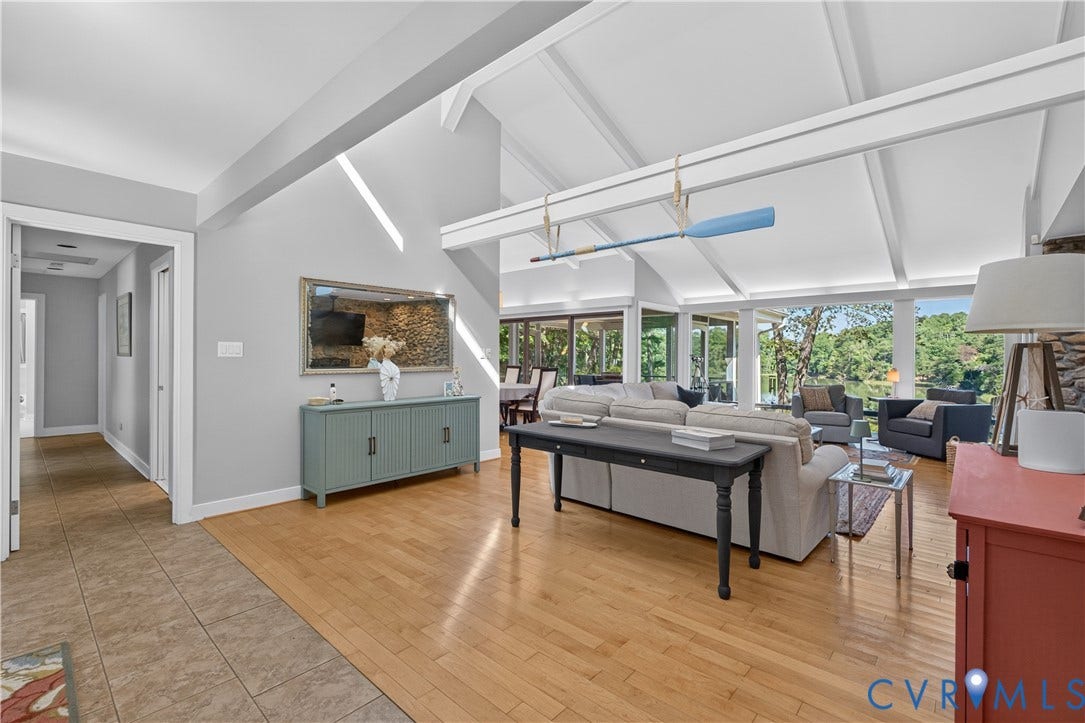 Vaulted ceiling with exposed beams, hardwood flooring, large windows, and a stone-faced accent wall