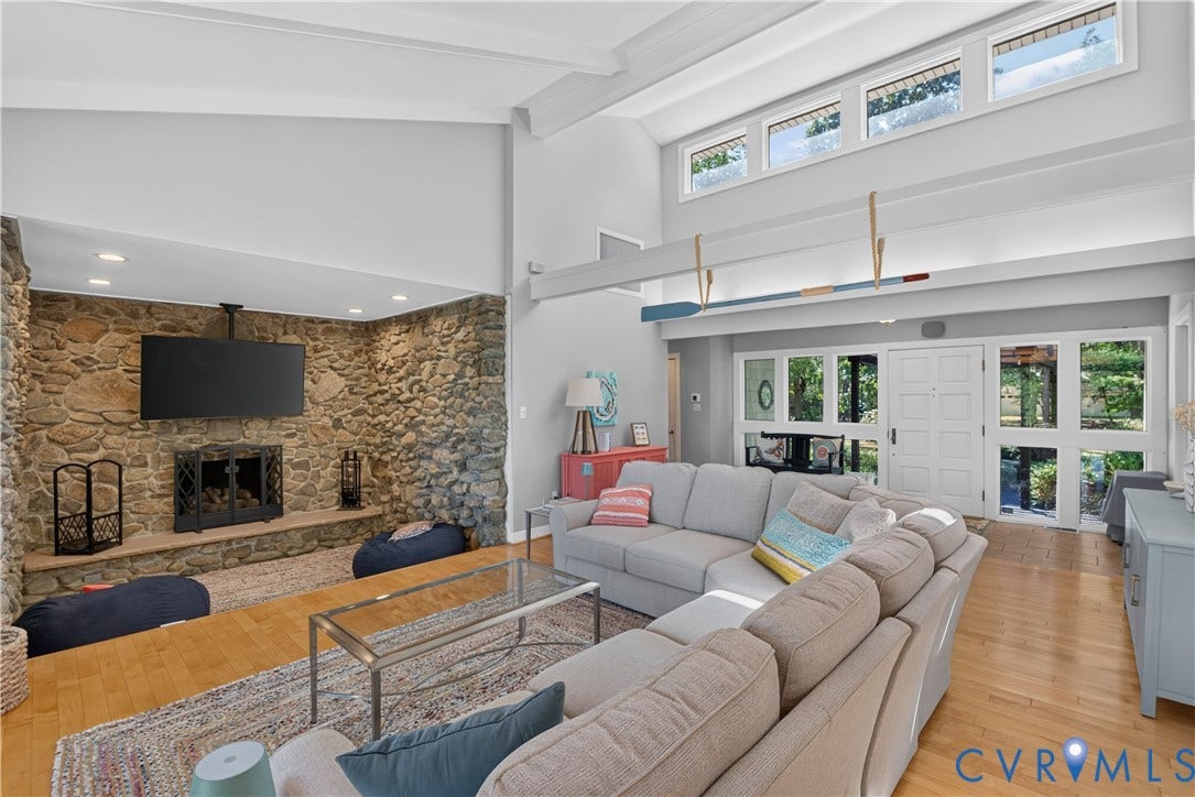 Vaulted ceilings with exposed beams, a stone-clad fireplace, light wood-finish flooring, and a double-door entry with surrounding windows