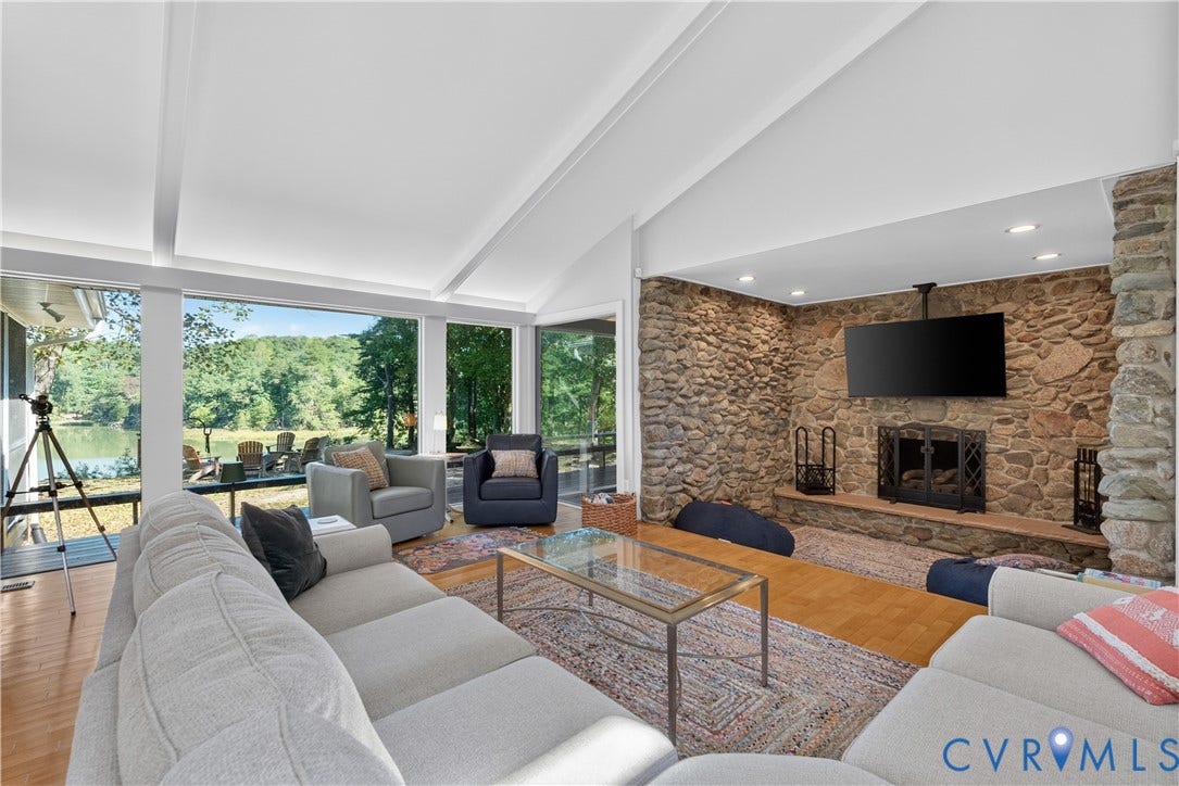Vaulted ceiling with exposed beams, stone-clad fireplace, recessed lighting, and expansive window wall with exterior scenery