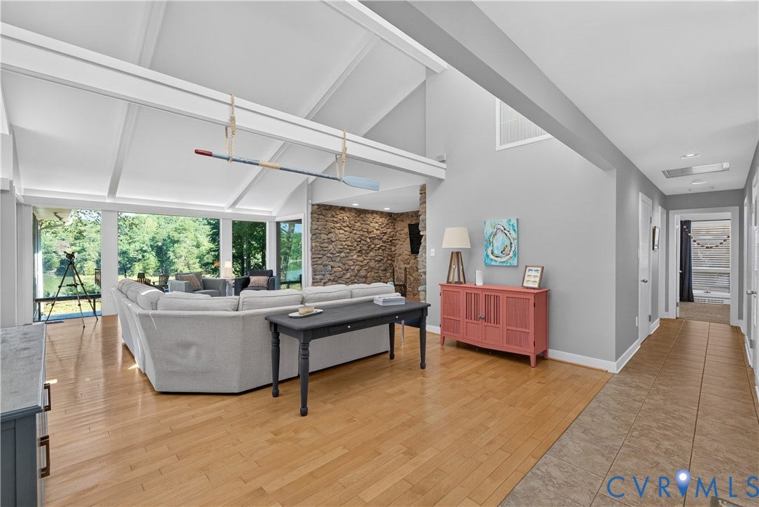Vaulted ceilings with exposed beams, stone accent wall, expansive window wall, light wood-finish flooring, and a tiled hallway