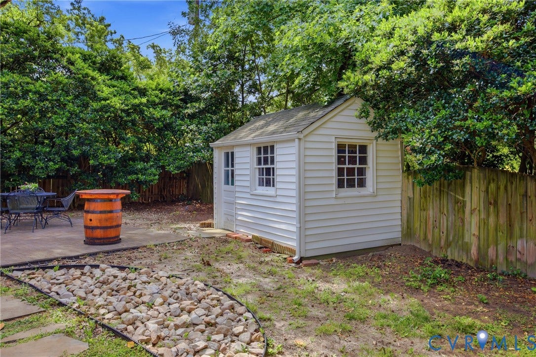White-sided shed with multiple pane windows, adjacent to a concrete patio, wood fencing, and a rock-lined garden bed