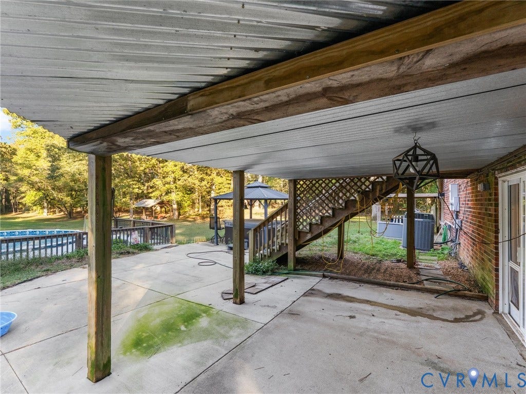 8251 Old Roxbury Road, Quinton, VA 23141 Covered concrete patio featuring wood structural supports, geometric metal pendant lighting, brick exterior wall, and wooden staircase with lattice railing