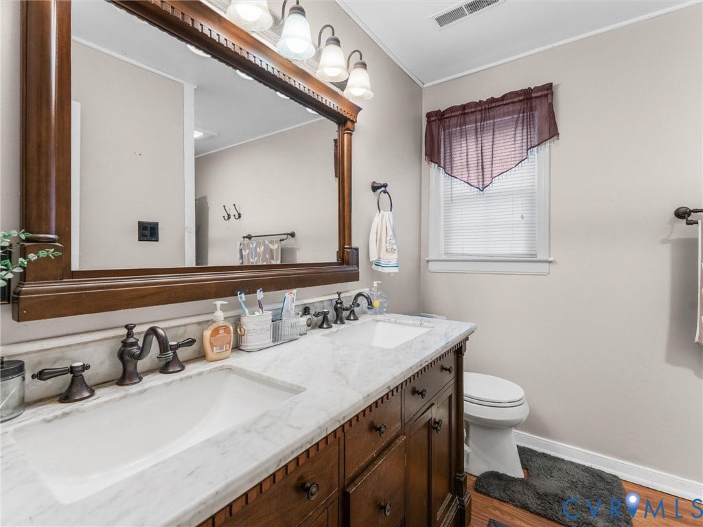 8251 Old Roxbury Road, Quinton, VA 23141 Double vanity with stone-surfaced countertop, integrated dual sinks, dark wood cabinetry, and an expansive framed wall mirror with integrated multi-bulb lighting