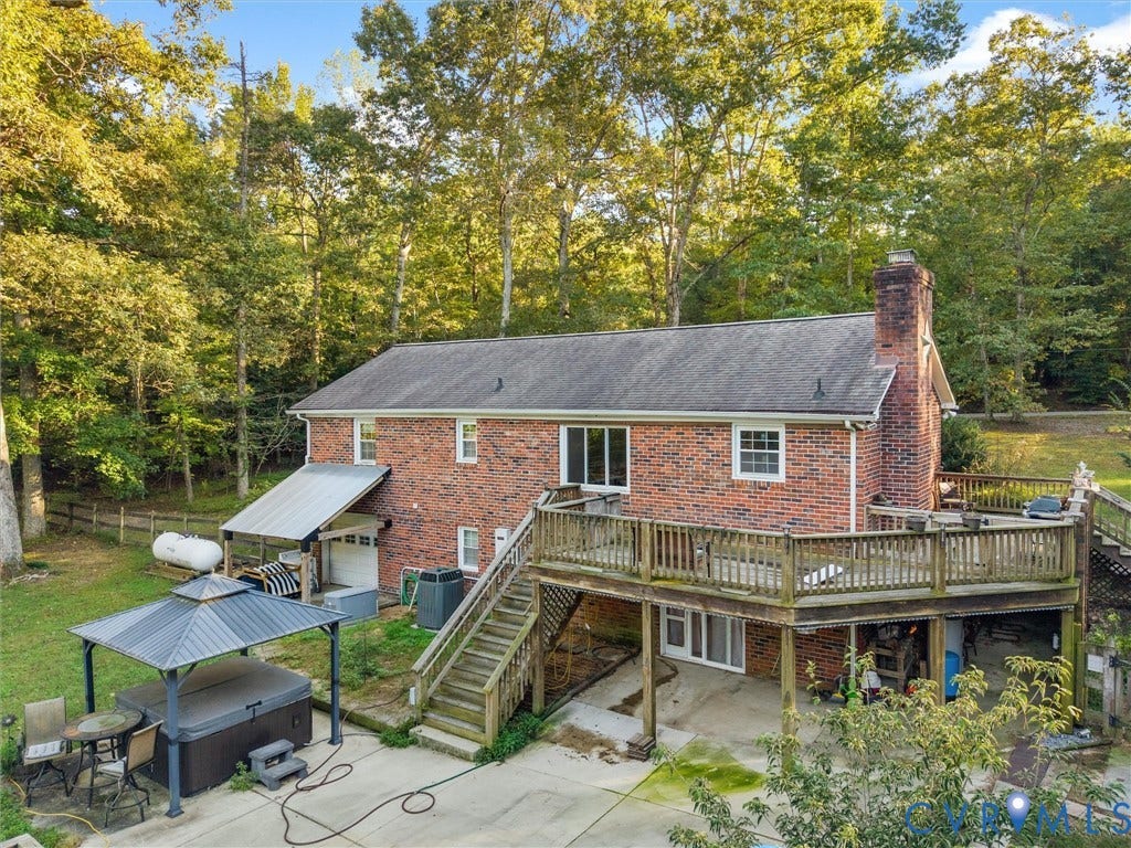 8251 Old Roxbury Road, Quinton, VA 23141 Brick exterior featuring an elevated wood deck, a prominent masonry chimney, a lower-level concrete patio, and a covered side storage area