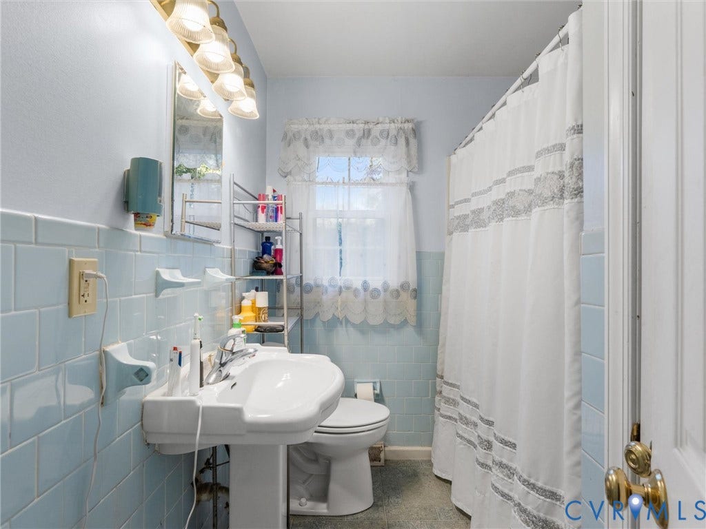 8251 Old Roxbury Road, Quinton, VA 23141 Bathroom featuring light blue tiled walls, white pedestal sink with polished chrome faucet, multi-bulb wall-mounted vanity lighting, and a window with a pleated valance
