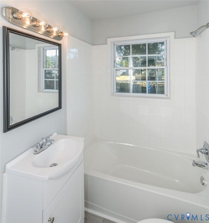 White subway tile bath surround, chrome shower fixture, single vanity with integrated sink, rectangular framed mirror, and wood-finish flooring