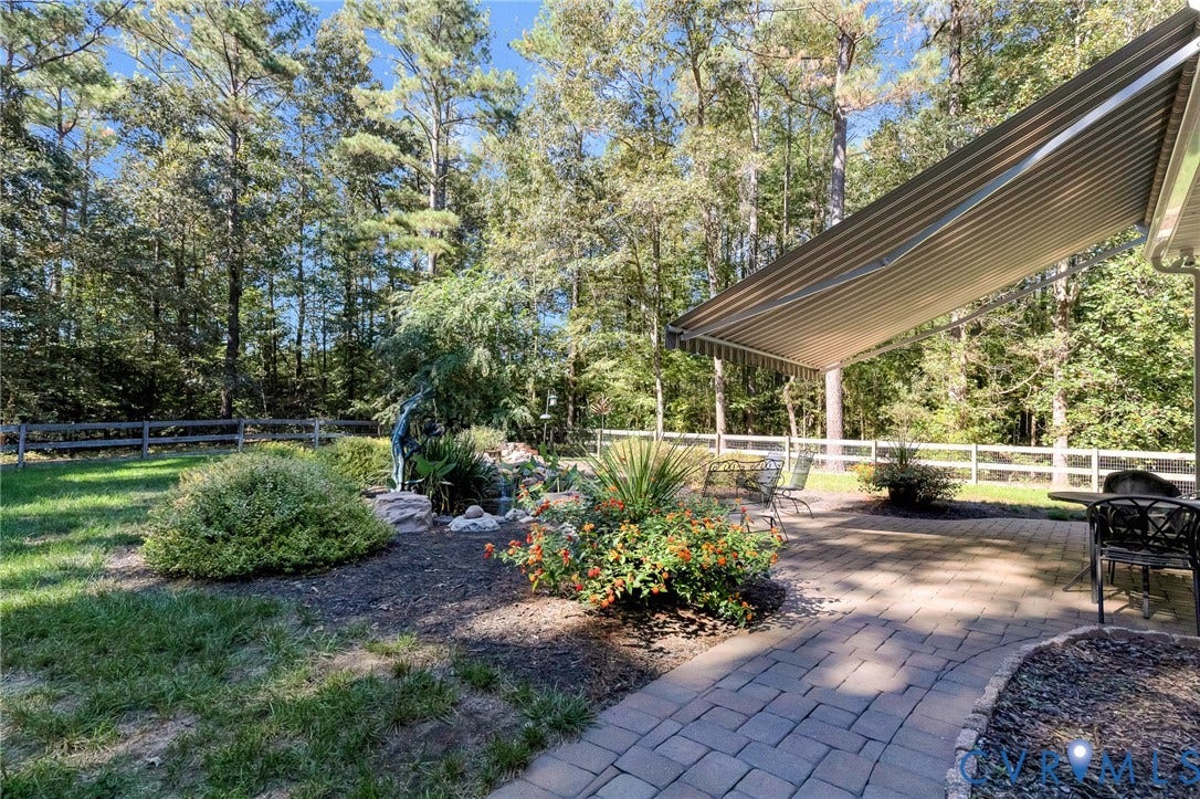 360 Laurel Drive, Aylett, VA 23009 Expansive paver patio with a retractable awning, surrounded by mature trees, a manicured lawn, and a decorative wooden fence