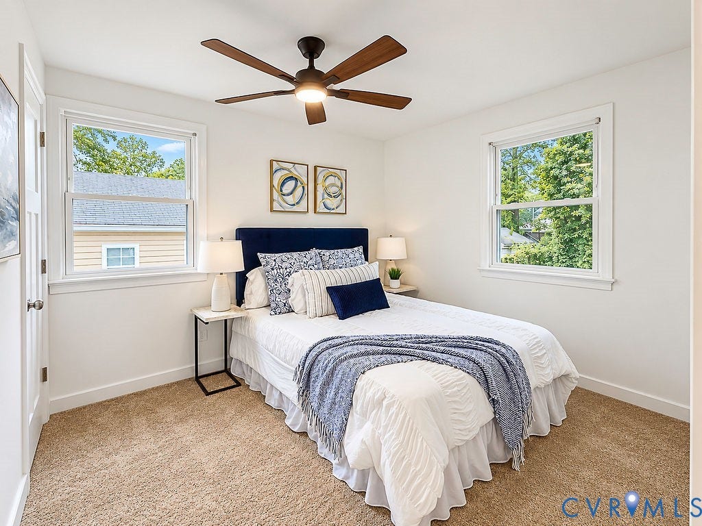 Neutral wall paint, plush carpet flooring, two double-hung windows, and a ceiling fan with integrated lighting