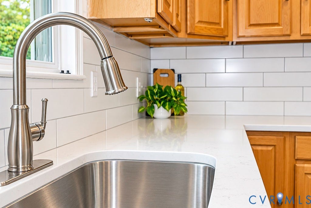 Stainless steel undermount sink with a gooseneck faucet, white subway tile backsplash, light-toned countertops, and wood cabinetry