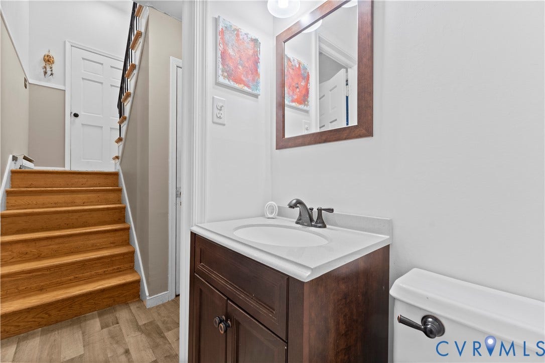 109 Webster Road, Sandston, VA 23150 Wood-finish flooring, staircase with black metal balusters, dark wood vanity with white countertop, wall-mounted mirror with wood frame, and white paneled door