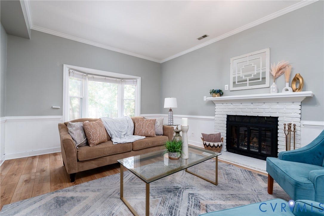 Living area with wood-finish flooring, a white brick fireplace with a mantel, and a bay window.