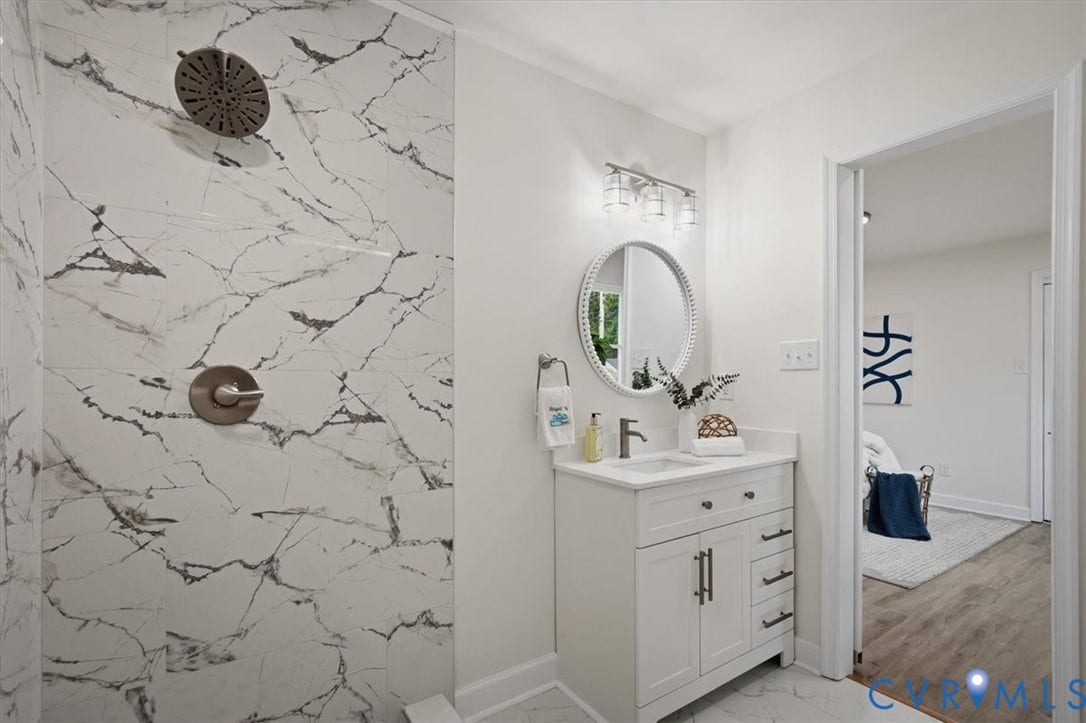 Walk-in shower with marble-look tile, brushed nickel shower fixtures, white vanity with integrated sink, modern vanity light fixture, and round decorative mirror