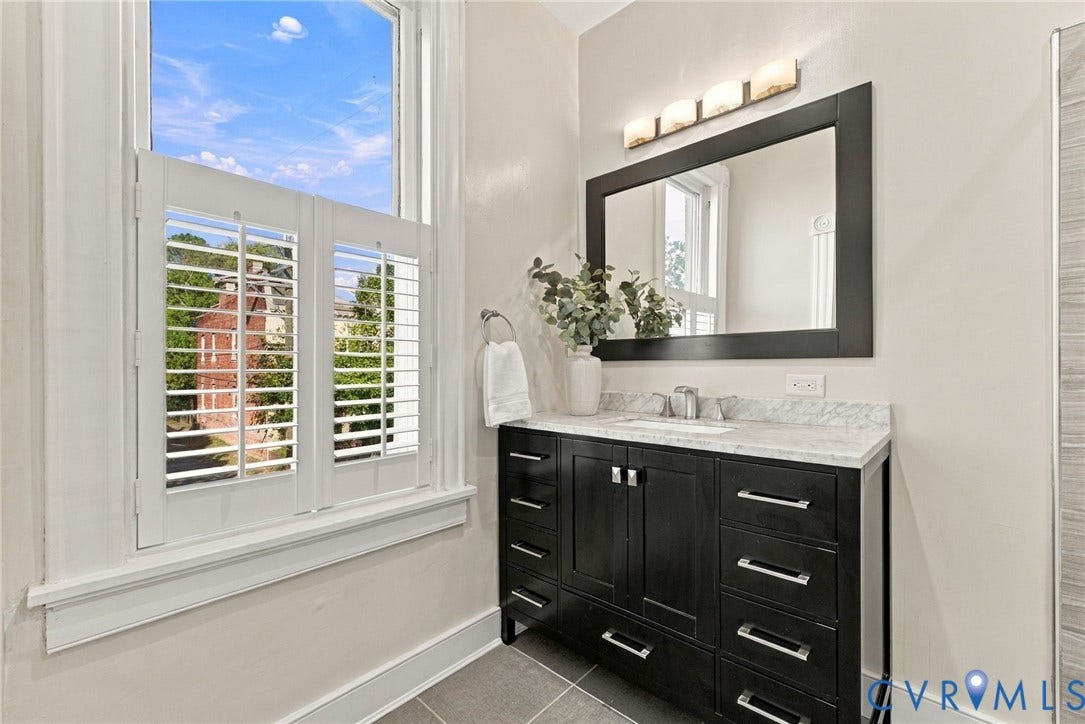 107 N Allen Avenue, The Fan, Richmond, VA 23220 Dark wood vanity featuring a stone countertop with integrated sink, rectangular mirror with dark frame, four-light wall fixture, and white plantation shutters