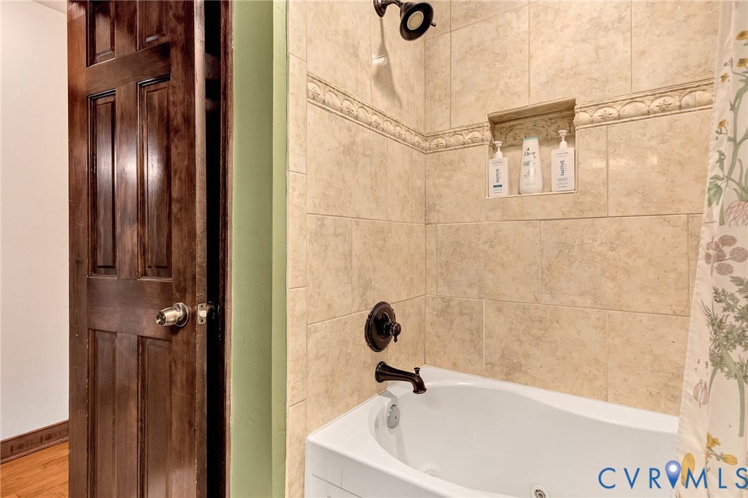 11410 Driscoll Road, Chester, VA 23831 Soaking tub with a matte black faucet and shower head, surrounded by neutral-toned stone-finish wall tile, an integrated recessed soap niche, and a decorative tile border