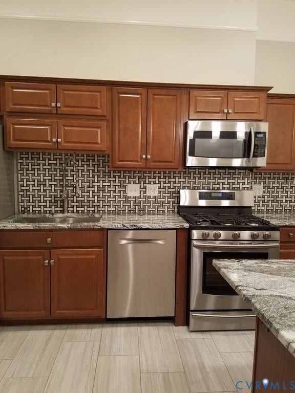 Wood-finish cabinetry, stainless steel appliances, mosaic tile backsplash, stone-finish countertops, and light-toned tile flooring