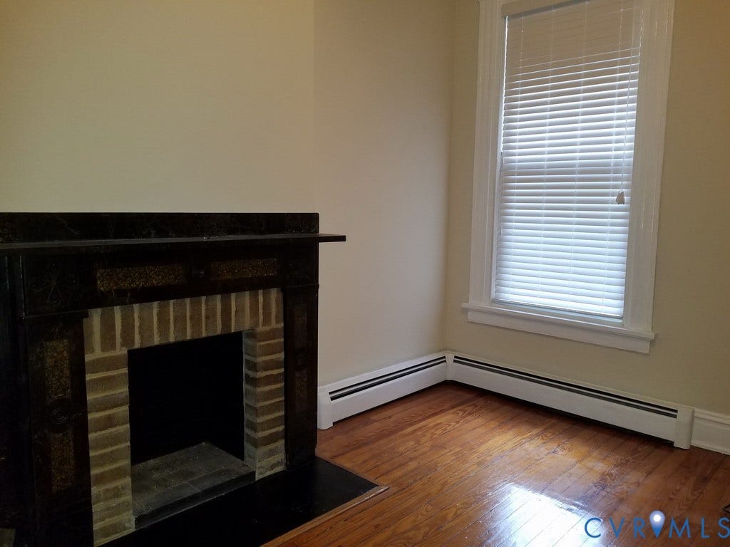 Hardwood flooring, fireplace with brick surround, dark mantel, single window with blinds, and baseboard heating