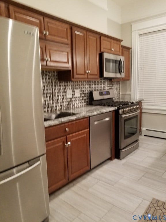 Kitchen featuring stainless steel appliances, wood cabinetry, stone countertops, a tile backsplash, and light-toned tile flooring