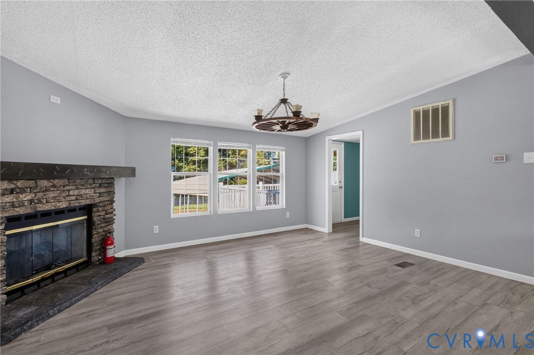 8211 Brills Road, Dinwiddie, VA 23872 Stone-faced fireplace with a dark wood mantel, wood-finish flooring, multiple windows, and an overhead chandelier with fan blades