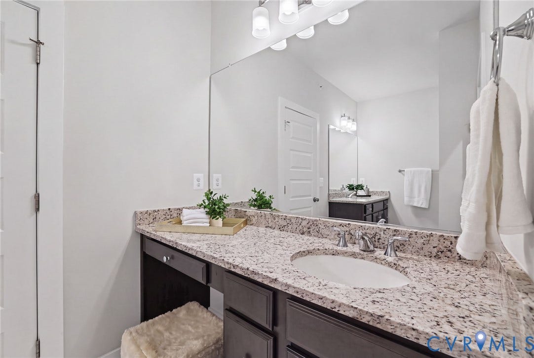597 Weaver Brook Walk, #B, Richmond, VA 23233 Dark-toned cabinetry featuring an integrated vanity, neutral stone-finish countertop, under-mount sink with dual-handle polished chrome faucet, and full-width wall-mounted mirror