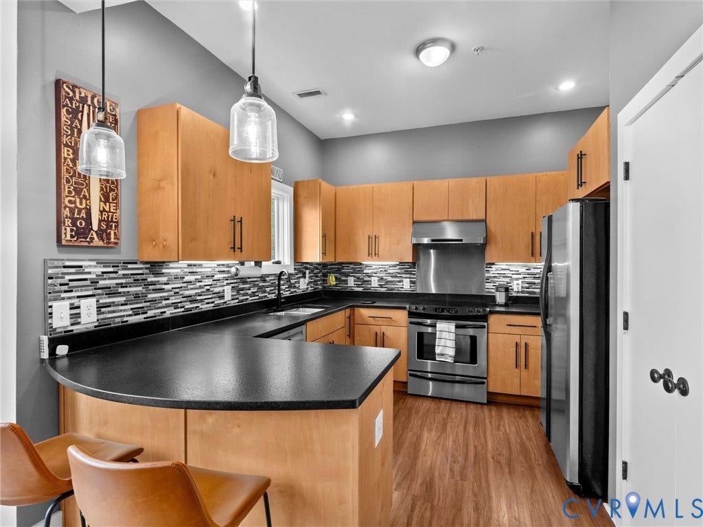 Kitchen featuring wood-finish cabinetry, black countertops, a mosaic tile backsplash, stainless steel appliances, and pendant lighting