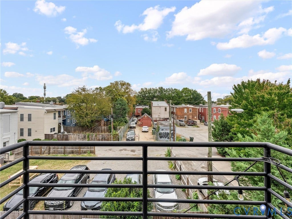 Balcony with black metal railing overlooking a paved parking area and residential structures