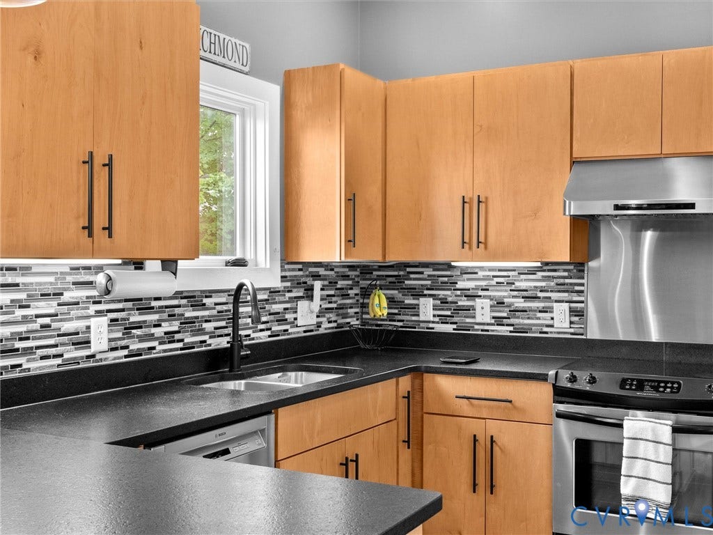 Light wood-finish cabinetry, dark countertops, mosaic tile backsplash, stainless steel appliances, and a black-finish sink faucet