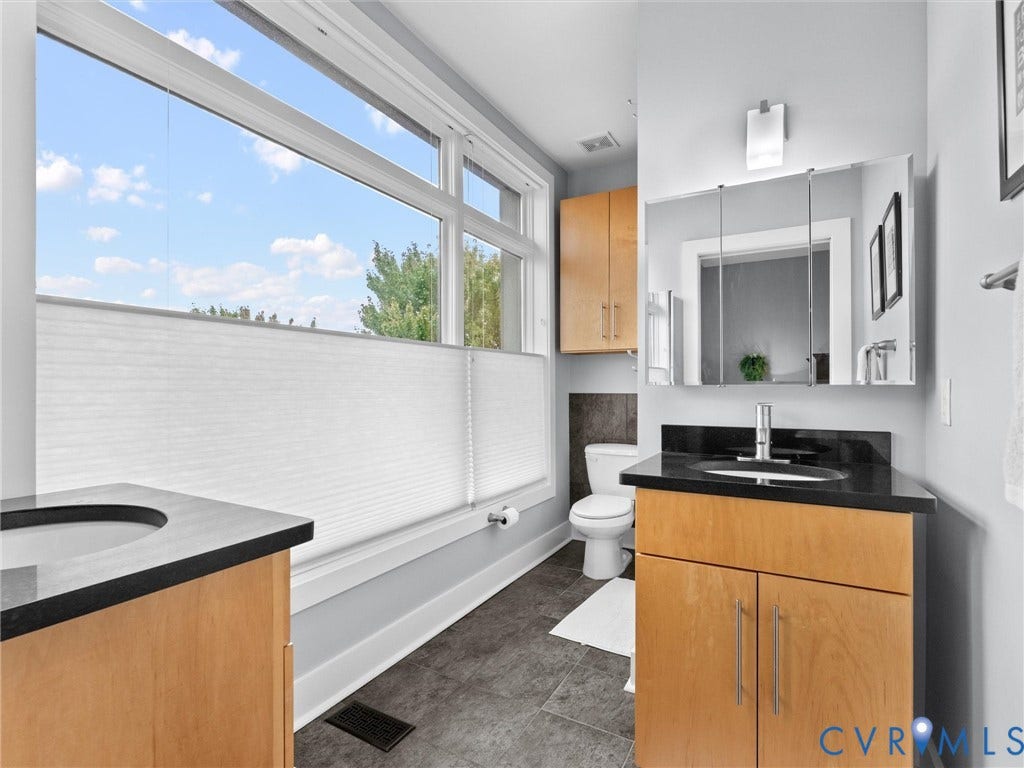 Dual vanities with dark countertops, built-in medicine cabinet, wood-finish cabinetry, and large windows with blinds