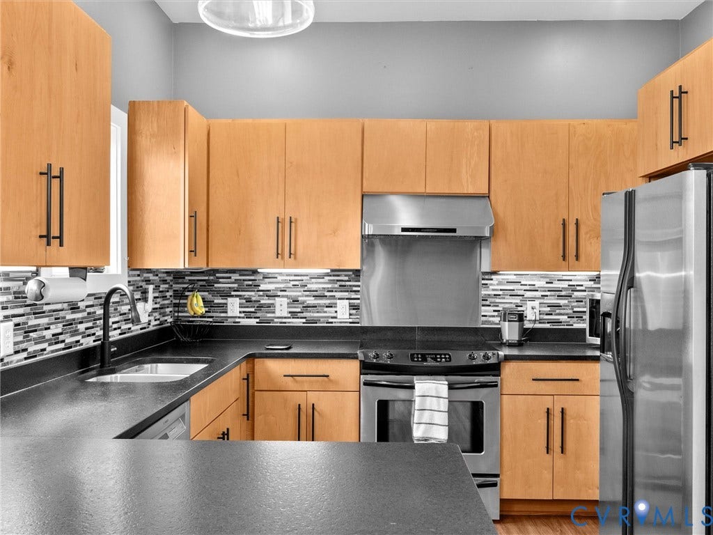 Kitchen featuring light wood cabinetry, matte black hardware, stainless steel appliances, black countertops, and a mosaic tile backsplash